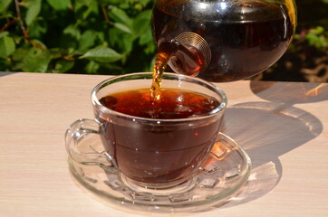 A girl pours tea from a transparent teapot into a transparent cup against a background of green foliage Good morning, outdoor cafe, energy boost. Tea being poured into glass tea