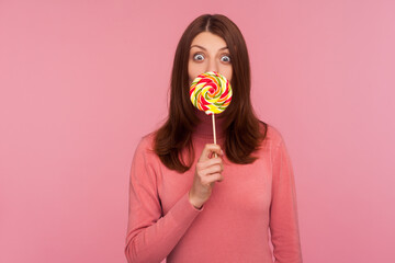 Surprised brunette woman in pink sweater licking lollipop and looking at camera with amazed expression. Indoor studio shot isolated on pink background