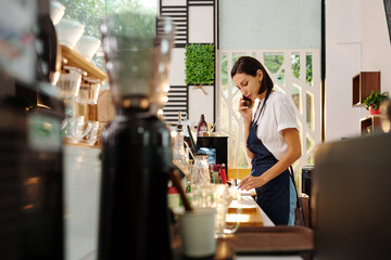 Pretty young female coffeeshop manager talking on phone with customer and writing down his order