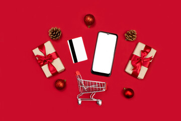 Smartphone mockup. Gifts, credit card, smartphone, fir cones, red decoration, shopping cart flat lay on red background top view, copy space. Online christmas shopping. 2021 new year, xmas holidays