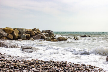 Stormy sea. Powerful big foam waves beat against big beautiful rocks, rocks, breakwaters. A scattering of spray. Cloudy weather with bright light.