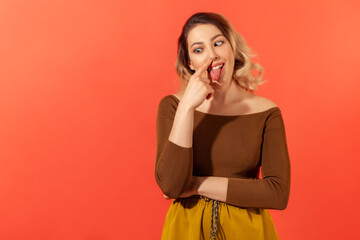 Fototapeta premium Portrait of funny woman in casual clothes touching her nose with forefinger, crossed eyes and tongue out. Funny and foolish concept. Indoor studio shot isolated on red background