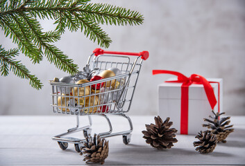 Christmas balls are in the shopping cart next to the gift box and cones on light wooden background