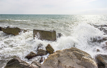Stormy sea. Powerful large foam waves beat against rocks and rocks. A scattering of spray. Cloudy weather with bright light.