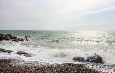 Stormy sea. Gray-blue, cloudy. Foam waves beat on the shore, rocks, breakwaters. Cloudy weather. Sunlight reflects in the water and flickers.