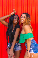 Lifestyle of friends, Caucasian blonde girl and black girl with big braid on a red background. Modern photography of urban girls in the city, posed hugging each other