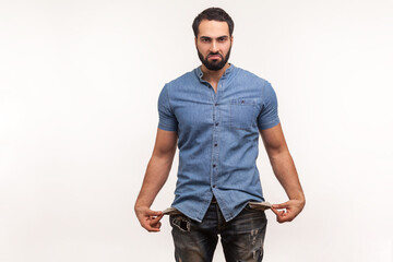 Poor upset man with beard in leaky jeans and blue shirt showing empty pockets looking at camera with sadness, bankruptcy, financial problems. Indoor studio shot isolated on white background