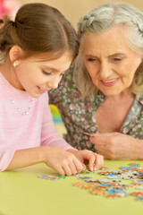 Fototapeta premium Cute little granddaughter and grandmother collecting puzzles together at home