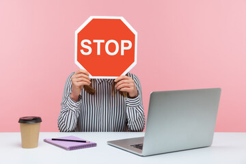 Female office worker in striped shirt sitting at workplace hiding face behind red stop traffic sign...