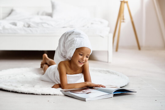 Little model girl wrapped in a towel lying on the floor on a carpet and reading a fashion magazine.