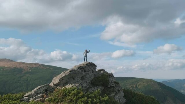 Happy man at top mountain celebrates climb to peak by jumping, dancing. Backdrop of mountains, blue sky, clouds. Caucasian man rejoices at achieved success in mountains. Success, victorious concept
