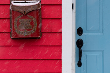 A bright red exterior wooden wall with a metal mailbox inscribed with North Pole Post. There's a blue door with a black handle and lock and a brass doorbell. There's white snow falling diagonally.  