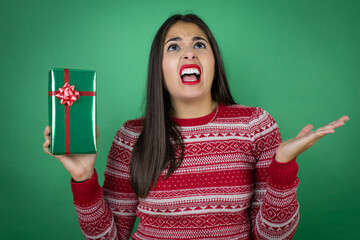 Young beautiful girl holding gift over isolated white background clueless and confused expression with arms and hands raised