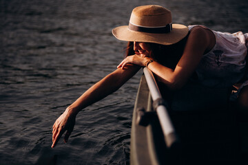 romantic girl in a straw hat sits in a boat and touches the water with her hand