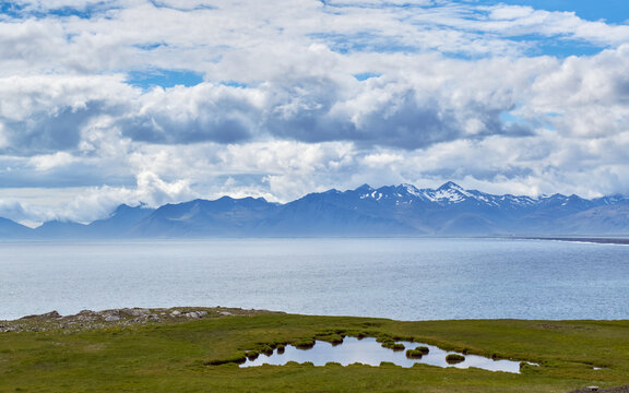 A small pond reflects the sky on a grassy plain on Hvalnes in eastern Iceland, with distant snow-capped mountains rising beyond the shoreline.