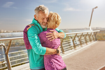 Fototapeta na wymiar Togetherness. Happy mature family couple in sportswear embracing after having workout in the city park on a sunny morning. Joyful senior couple standing together outdoors