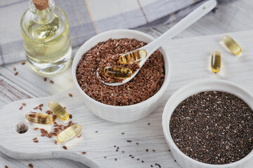 Brown flax seed or linseed and chia in small bowl and gelatin capsules with omega oil on a white wooden background.