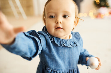 Portrait of a little baby girl dressed in a blue denim dress standing in a bright room.