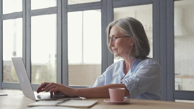 Senior older professional mature business woman using laptop computer sits at workplace desk. Happy mid aged employee 60s grey-haired businesswoman executive working typing on pc at home from office.