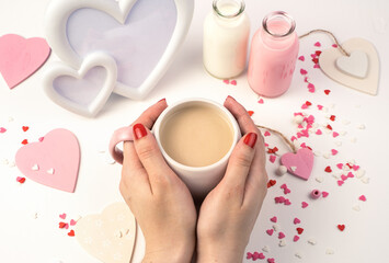 Hands of a woman holding a Cup of milk coffee on a background of hearts, a white frame and bottles of milk on a white background. Concept February 14.