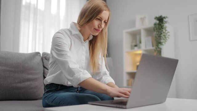 Confident woman in white shirt and blue jeans using wireless laptop for work while staying at home. Happy blonde sitting on couch and doing her job on distance.