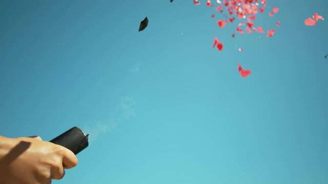 SLOW MOTION, CLOSE UP, COPY SPACE: Unrecognizable person fires a confetti gun at the clear blue sky. Pink and red heart shaped pieces of paper explode out of black barrel and high into the skies.