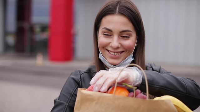 Groceries in female's hand by the car truck. Woman in protective mask and disposable gloves. Fresh fruits in eco paket. Food shopping during coronavirus Covid-19 quarantine. Woman take apple to feel