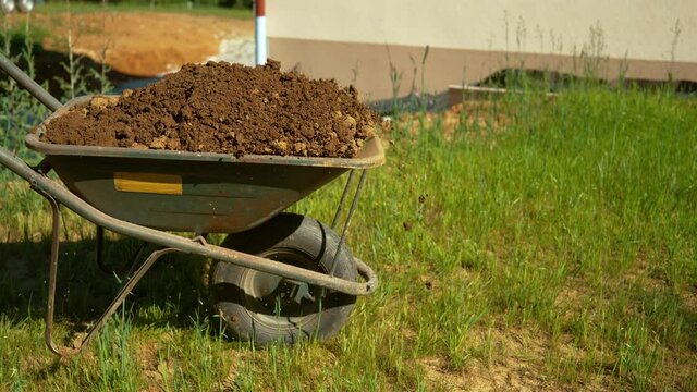 SLOW MOTION, CLOSE UP, DOF: Big metal wheelbarrow full of wet soil is pushed across the backyard. Unknown gardener scatters soil while transporting it in a barrow from one end of property to a garden.