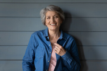 Head shot portrait smiling mature grey haired woman standing on grey wooden wall background, happy attractive senior older female looking at camera, posing for photo at home, retirement