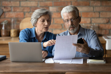 Serious mature couple checking financial documents, domestic bills, planning budget together, sitting at desk with laptop, senior man and woman counting taxes, discussing bank debt or loan payment