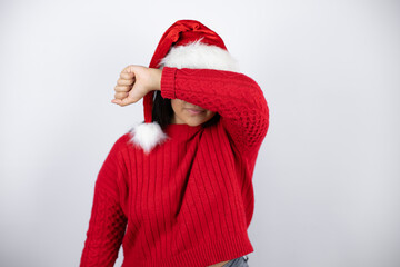 Young beautiful woman wearing a Santa hat over white background covering eyes with arm smiling cheerful and funny. Blind concept.