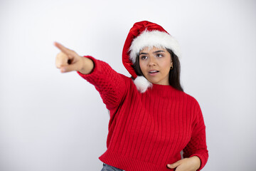 Fototapeta na wymiar Young beautiful woman wearing a Santa hat over white background pointing with finger surprised ahead, open mouth amazed expression, something on the front