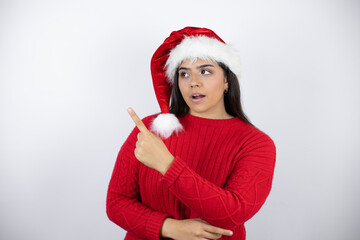 Young beautiful woman wearing a Santa hat over white background surprised and pointing with hand and finger to the side