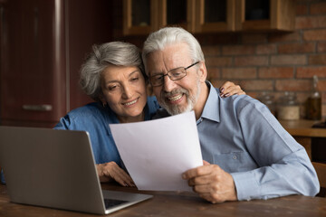 Smiling mature married couple reading letter with good news, using laptop, sitting at table in kitchen, happy grey haired wife hugging senior husband holding paper sheet, document, money refund