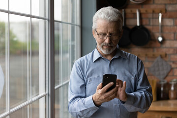 Close up satisfied mature man wearing glasses using phone, standing near window at home, cheerful senior male looking at smartphone screen, browsing apps, shopping or chatting with relatives online