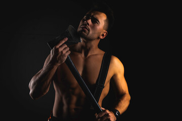 young handsome adult, muscular firefighter in uniform holding ax fire equipment in hands, pensive, isolated on dark background. Low key