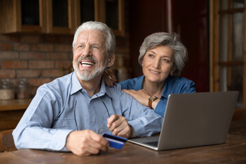 Happy dreamy mature couple hugging, sitting at table with laptop, overjoyed senior man holding plastic credit card, preparing to make secure internet payment, shopping online, elderly insurance