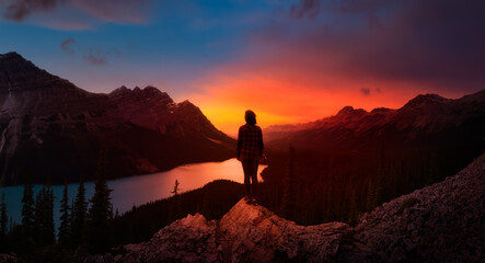 Adventurous girl standing on the edge of a cliff overlooking the beautiful Canadian Rockies and Peyto Lake. Dramatic, vibrant summer sunset. Taken in Banff National Park, Alberta, Canada.