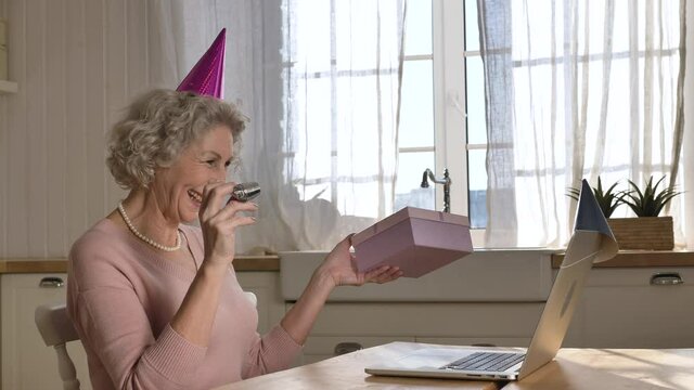 Happy retired woman with purple cone on head blows paper pipe holding birthday present box against laptop in kitchen closeup