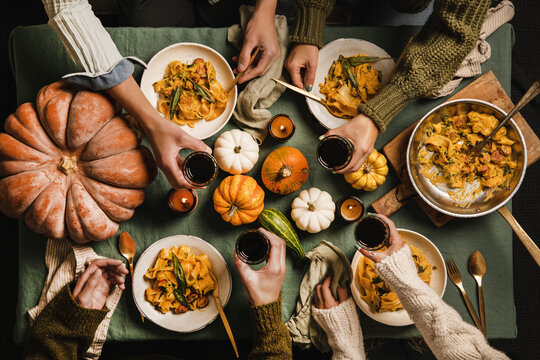Flat-lay of Autumn dinner for gathering or Thanksgiving Day celebration. Friends clinking glasses with red wine and eating butternut squash pasta with sausage and sage over linen tablecloth, top view