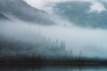Dense fog above tranquil mountain lake. Calm water and stony steep slope with coniferous trees among low clouds. Ghostly atmospheric view. Minimalist scenery in mysterious place at early foggy morning