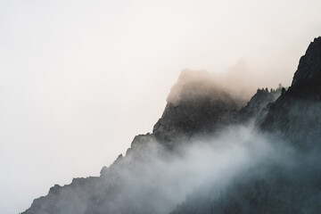 Fototapeta premium Ghostly atmospheric view to big cliff in cloudy sky. Low clouds among giant rocky mountains. Mysterious place at early foggy morning. Minimalist scenery with beautiful rockies. Dramatic bleak fog.