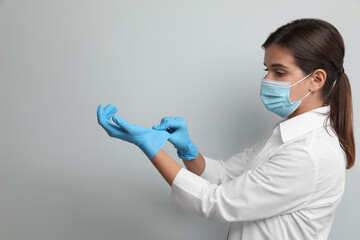 Doctor in protective mask putting on medical gloves against light grey background