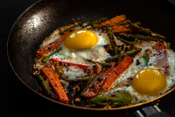 Very coarsely two fried eggs in a pan with large slices of carrots, bell peppers and asparagus with spices and sauce.