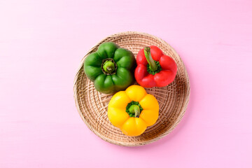 Green, red and yellow bell peppers on woven rattan plate with pink background, Top view