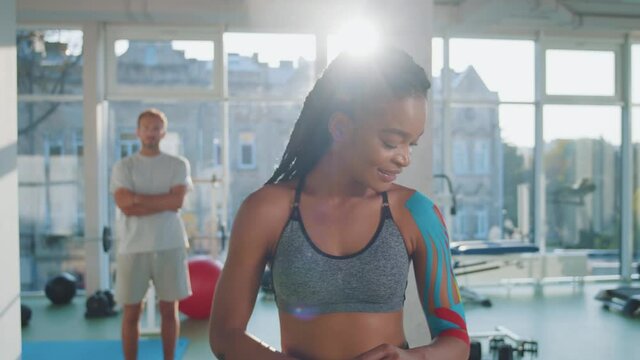 Young african american girl athlete standing in the middle of the gym with kinesio tapes on her left shoulder, stretches her hand and glad that she no longer hurts and the injury does not bother her