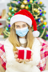 Portrait of a beautiful smiling woman in a Christmas hat holding a present, standing against the background of a Christmas tree. Woman in protective mask from virus, Christmas in quarantine