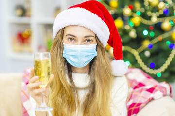 Portrait of a beautiful smiling woman in christmas hat  holding champagne glass while standing over cristmas tree background. The woman in the protective mask from the virus, Christmas in quarantine