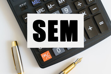On the table there is a pen, a calculator and a business card on which the text is written SEM. SEARCH ENGINE MARKETING