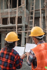 view from behind of male and female asian contractors standing wearing safety helmets holding the site plan building in the background of the unfinished building